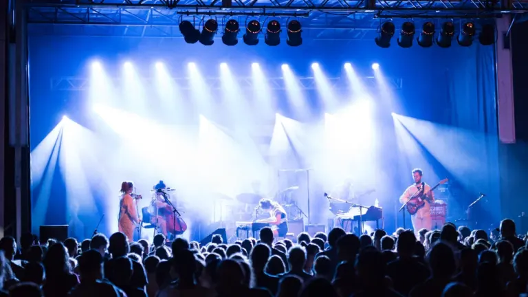 Muzikantų grupė koncertuoja scenoje su ryškiu apšvietimu priešais gausią žiūrovų auditoriją renginyje.