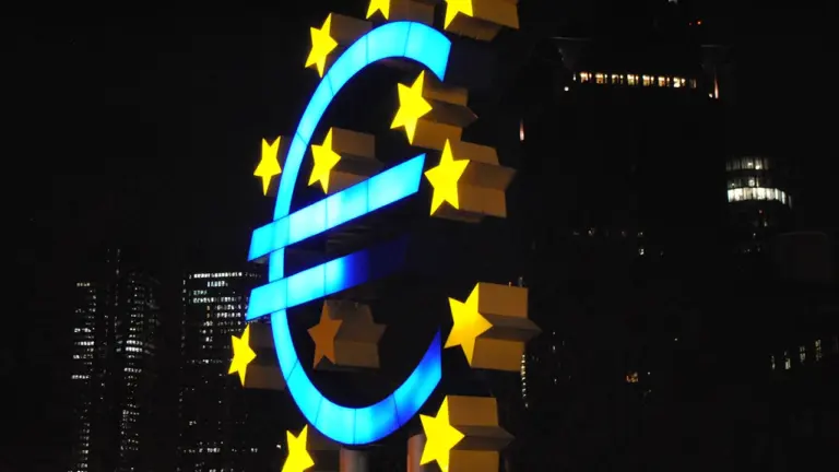 Šviečiantis Euro simbolis naktiniame mieste, simbolizuojantis Europos Centrinio Banko vykdomą pinigų politiką.