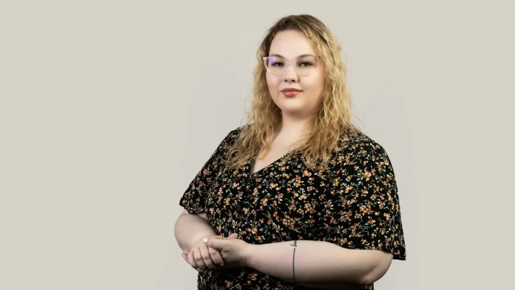 Šviesiaplaukė moteris su akiniais vilki gėlėtą suknelę ir stovi prieš pilką studijos foną.