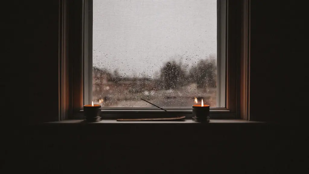 Jaukus vaizdas pro langą su lietumi, uždegtomis žvakėmis ir melancholiška nuotaika.