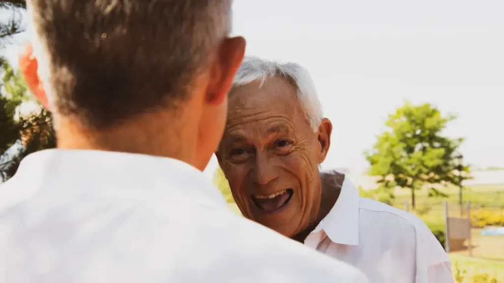 Vyresnio amžiaus vyras besišypsantis pokalbio su kitu žmogumi metu lauke.